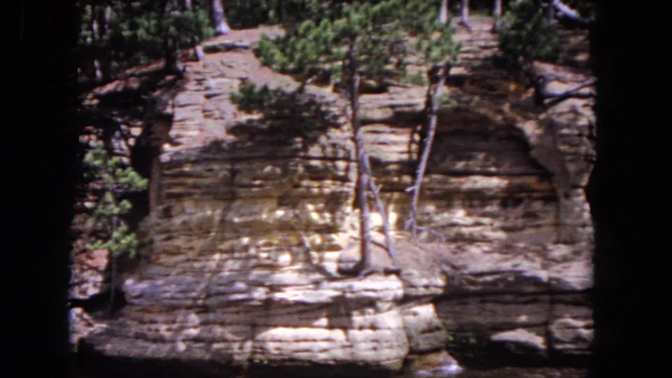 1964 Evanston 8mm view of a riverbank bluff with horizontal rock layers, exposed roots, evergreens, and water below.