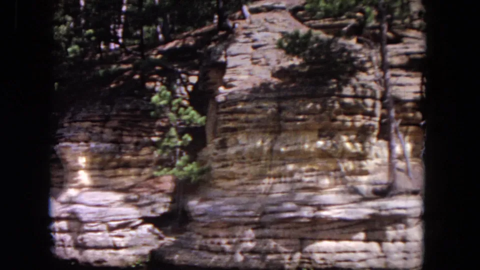 1964 8mm frame showing the pan across Evanston riverbank rock, with a small evergreen foregrounded against weathered ledges.
