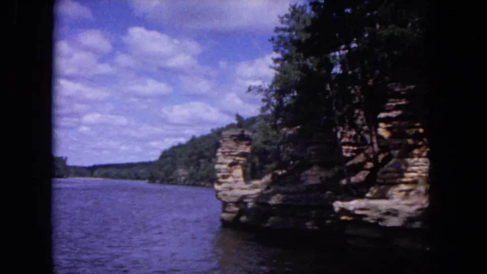 Wide 1964 home-movie frame from a boat, showing river water, a wooded bluff, layered rock, and a blue clouded sky.