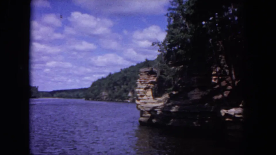 The boat looks past a rocky, tree-covered bluff toward open river water under a bright 1964 summer sky.