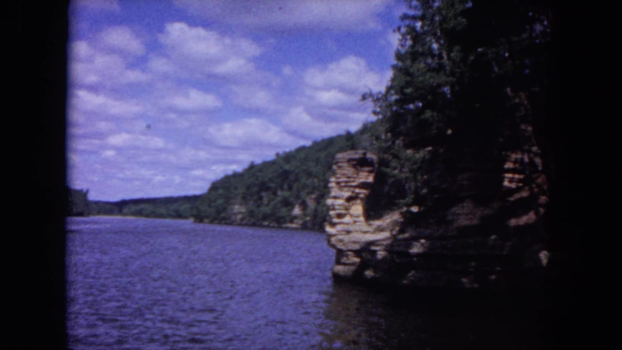 Lake bordered by wooded shoreline and layered rock cliffs under a blue sky.