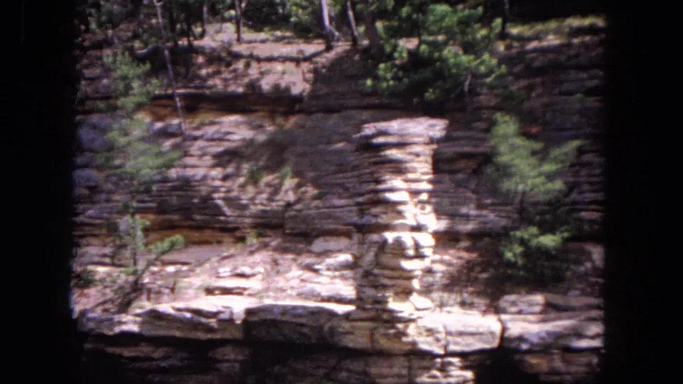 1964 home-movie frame of layered lakeside rock, small evergreens and a shallow dark opening in the cliff face.