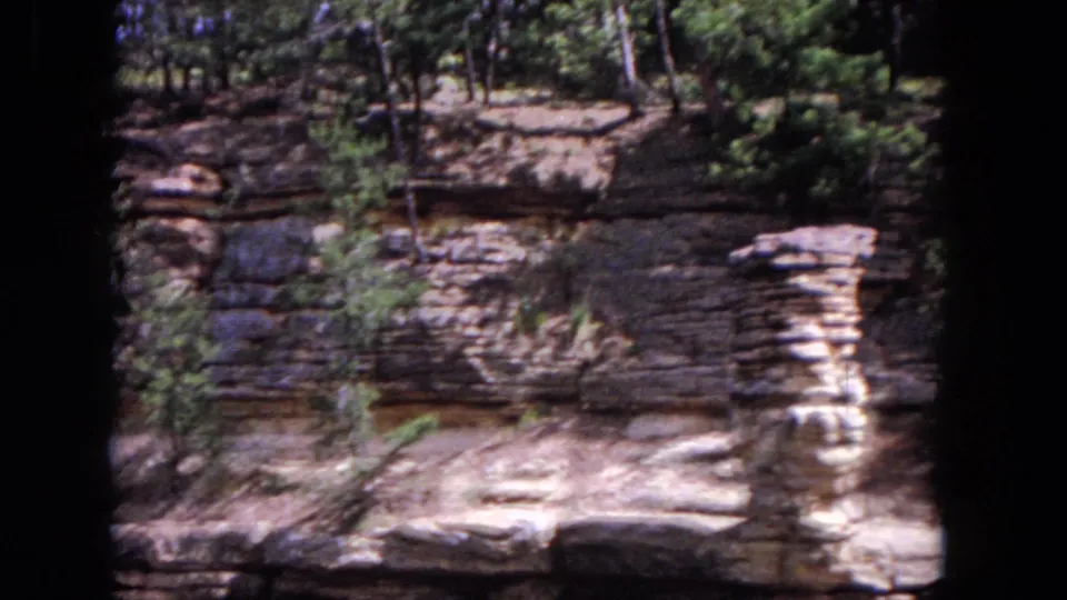 Final 8mm frame holds on the textured Evanston lakeside cliff, with trees above and pale rock ledges in sun.