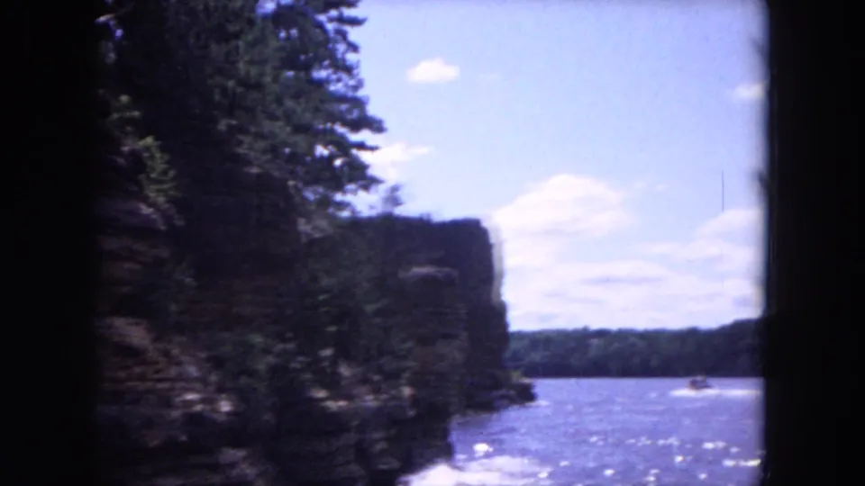 Color 8mm frame showing a shaded rock or wooded bluff beside bright water, within the 1964 Evanston, Illinois clip context.