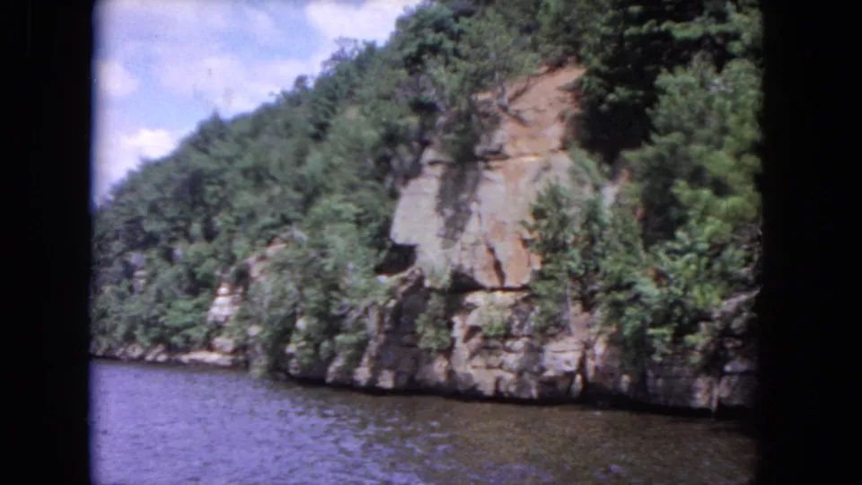 1964 8mm river view with layered rock, heavy greenery, and water filling the foreground from a moving vantage point.