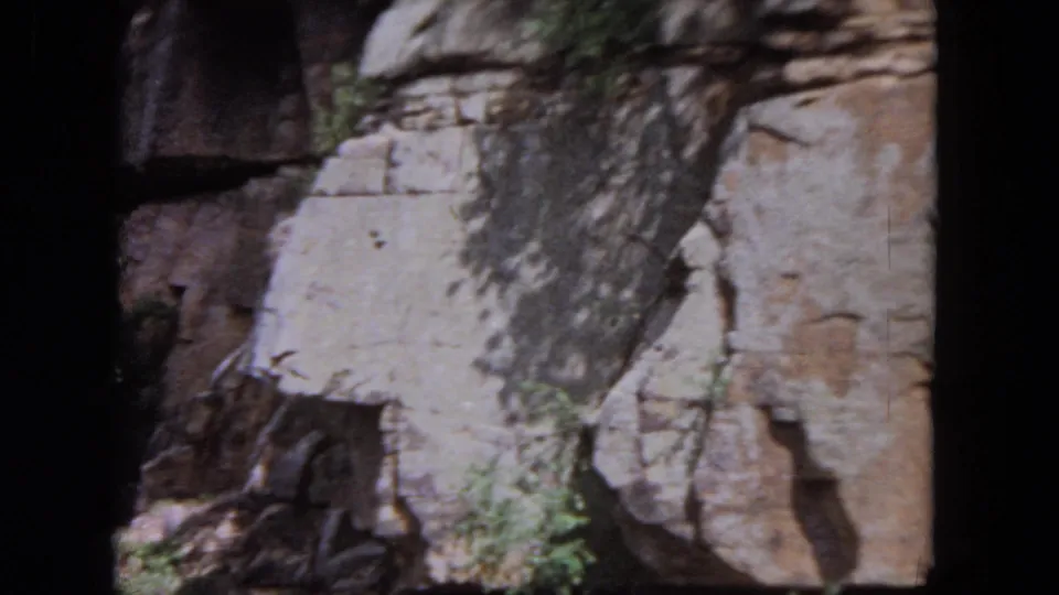 Closer 8mm view of a tan cliff face with cracks, small plants, and shadowed film-frame edges during the river pass.
