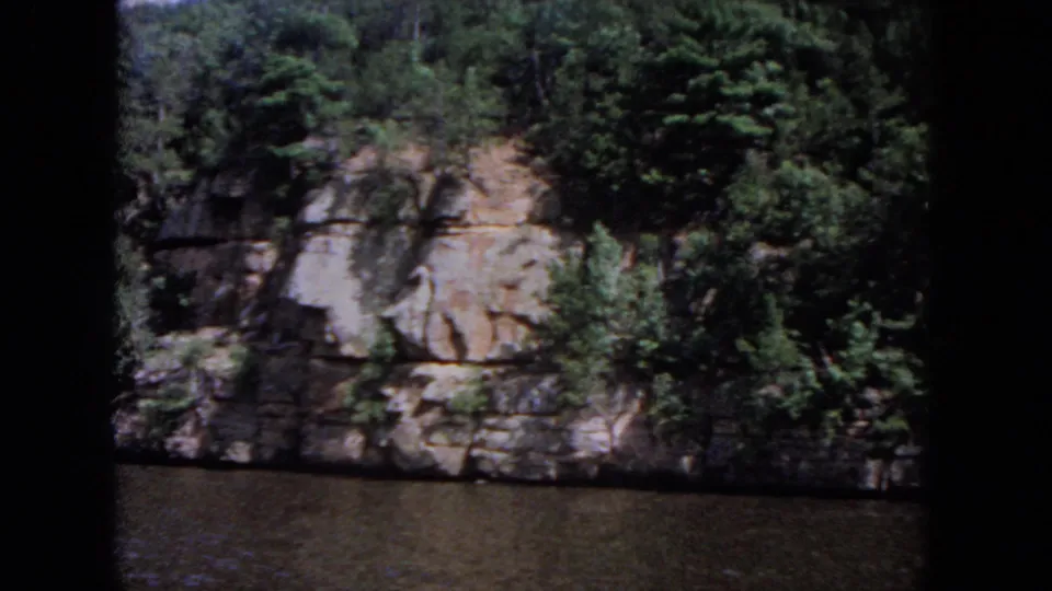 A wooded rock face rises above calm water in cataloged 1964 Evanston home-movie footage.
