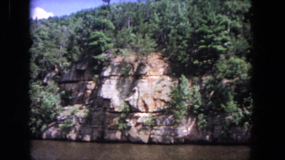 The 8mm frame shows a tree-covered rock bank above a slow river, with no visible people or structures.
