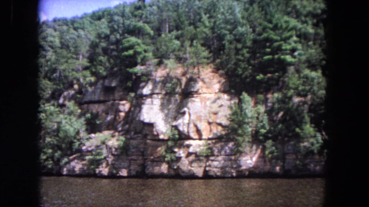 Forested rock bluff above dark river water with rounded 8mm frame edges.