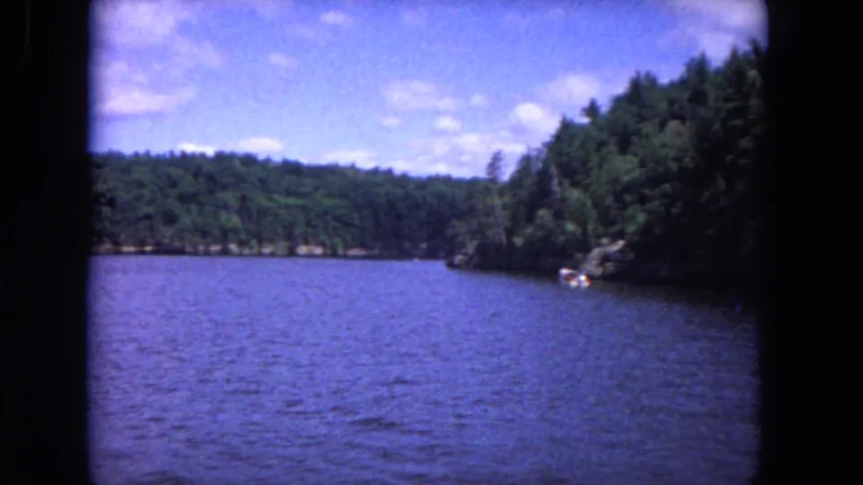 Wide 1964 8mm river view looking between wooded banks, with distant rocky shoreline, blue water, and a partly cloudy sky.