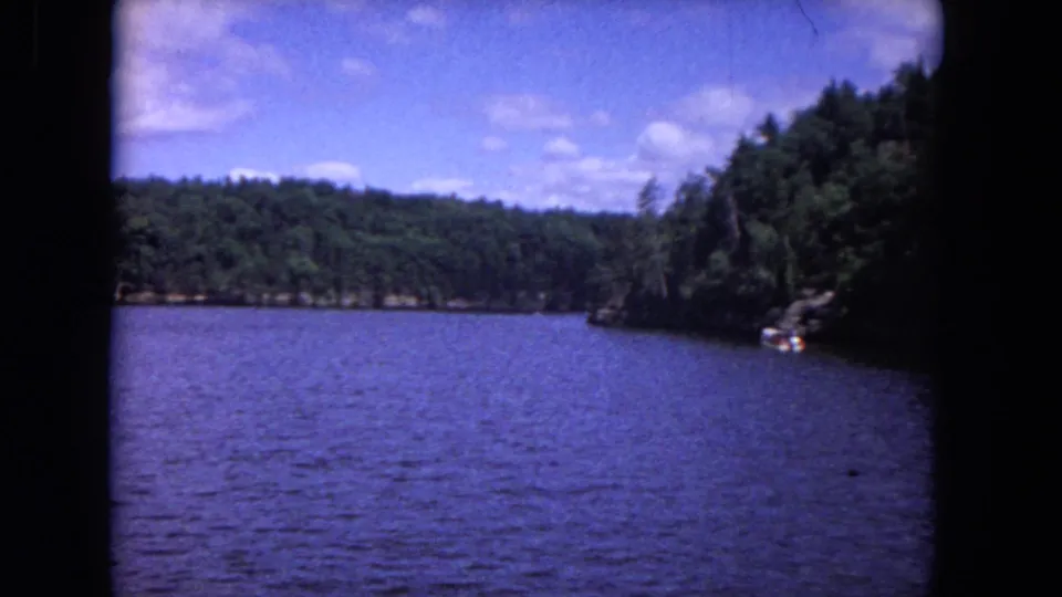 1964 8mm home-movie frame showing rippled river water, forested banks, and a shaded rocky edge from a moving boat-like viewpoint.