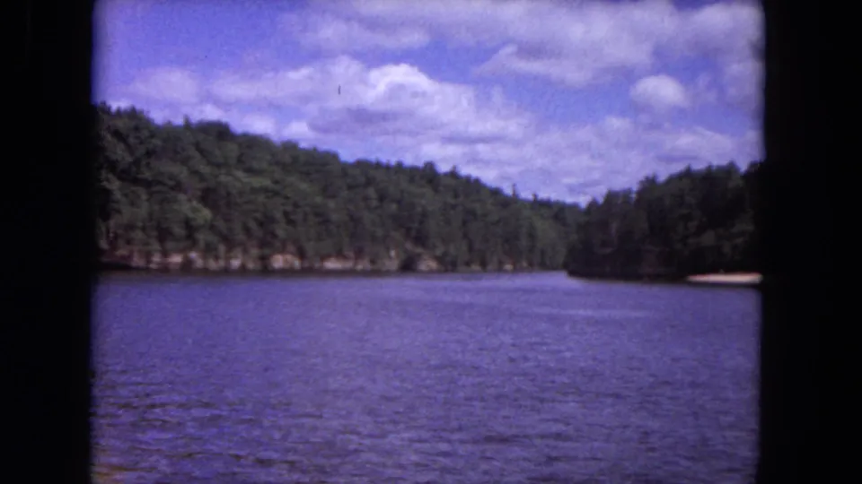 Final 1964 8mm frame of a broad river bend bordered by tree-covered bluffs, open water, and a bright clouded sky.