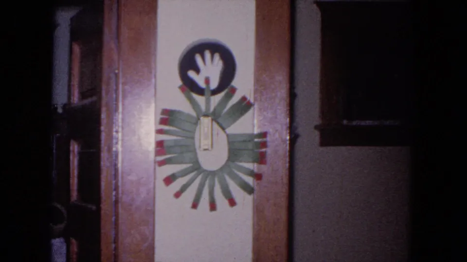 1964 Evanston home-movie frame showing a handmade child's handprint craft mounted on a wooden door or wall panel.