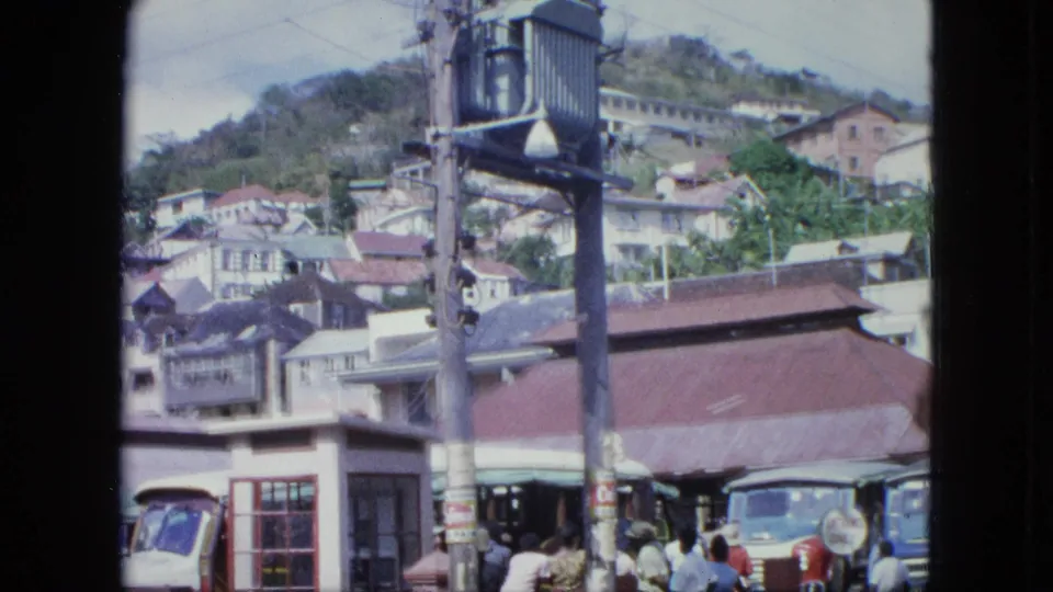 Archival 8mm view of a hillside town street, with clustered buildings, a utility pole, old vehicles, and people near the road.
