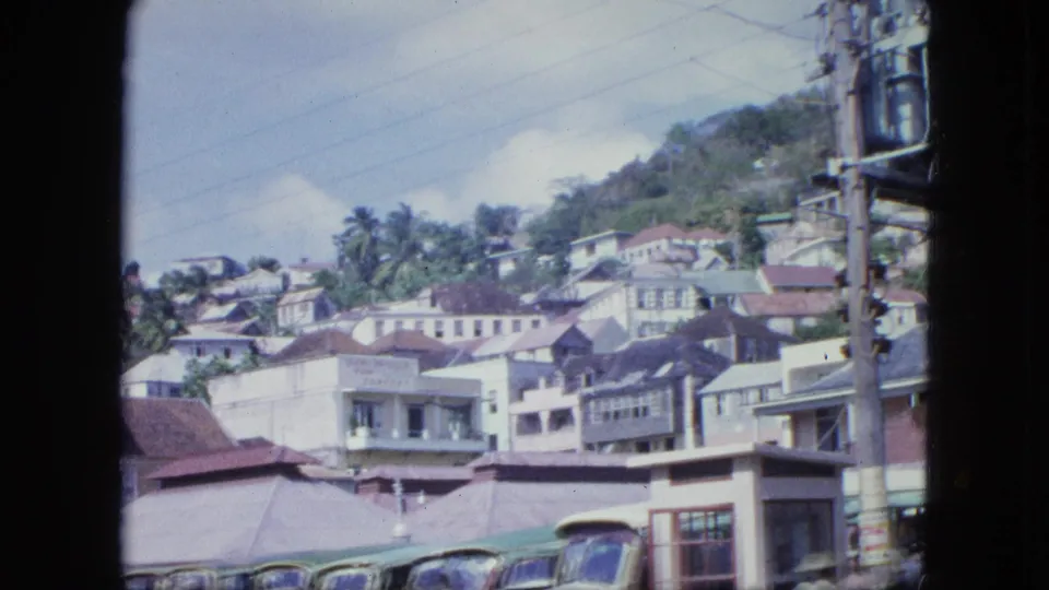 The frame looks across a compact hillside town with pale buildings, tiled roofs, palm trees, a utility pole, and vehicles below.