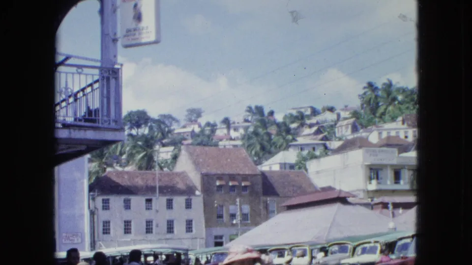 The view shifts across hillside buildings and palms, with a balcony or railing at left and parked vehicles along the lower street.