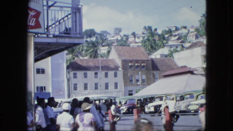 Adults in light clothing stand near a row of vintage cars, a covered stall, and hillside buildings in a busy street scene.