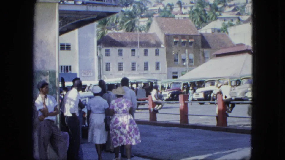 Adults gather in the shaded foreground while vintage cars, bollards, a covered structure, and hillside buildings fill the street beyond.