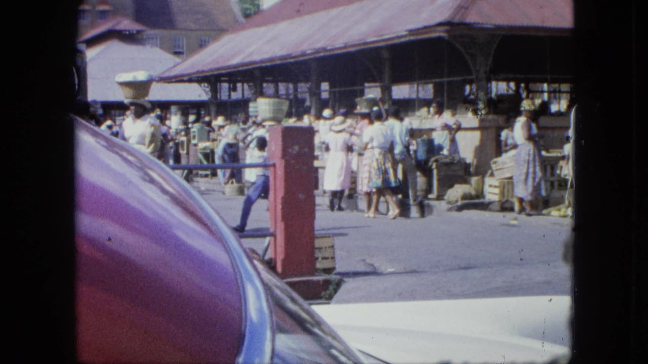 1964 Grenada market scene with shoppers under a covered structure, viewed over a purple-red classic car hood.