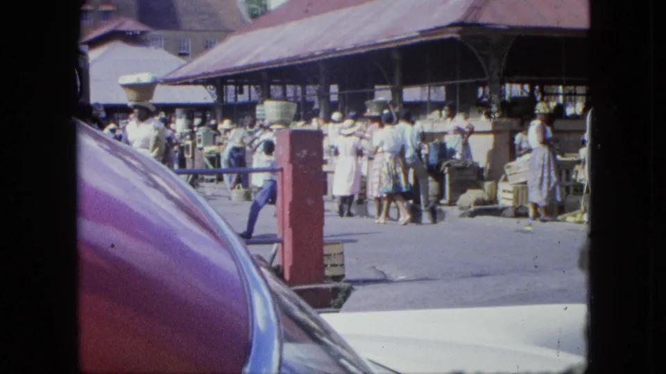 1964 Grenada market scene with shoppers under a covered structure, viewed over a purple-red classic car hood.