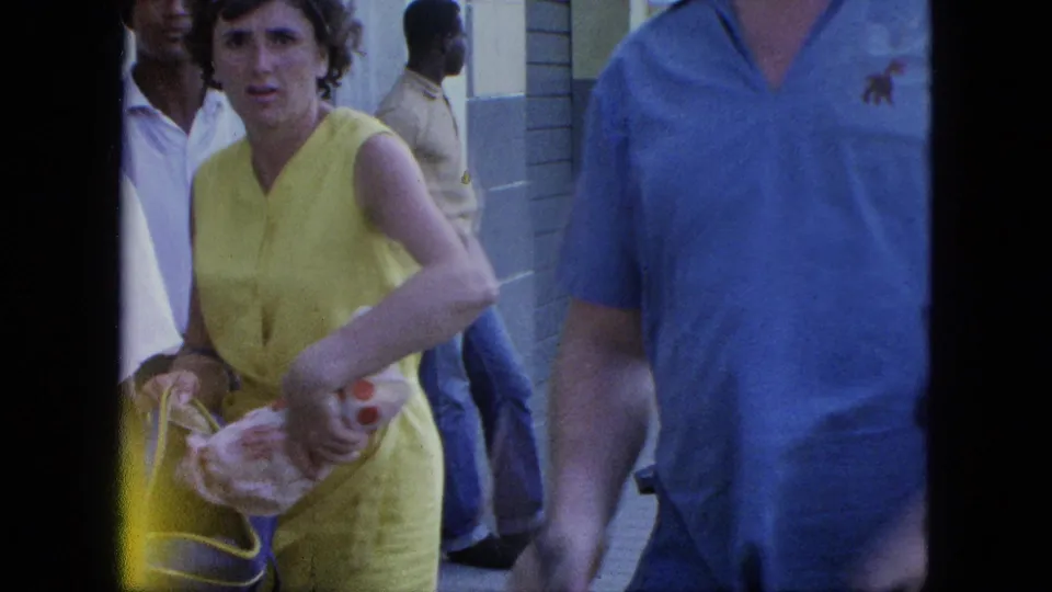 Close street-level home-movie frame of pedestrians, including a woman in a yellow dress carrying items near a blue-painted wall.
