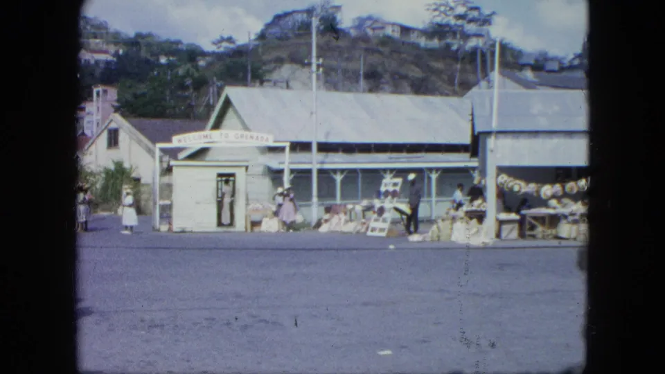 Wide 1964 Grenada street view with a Welcome to Grenada sign, roadside buildings, pedestrians, and goods displayed outside.