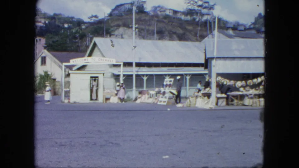 1960s Grenada home-movie frame showing a roadside market building with stalls, people, a visible sign, and hillside background.