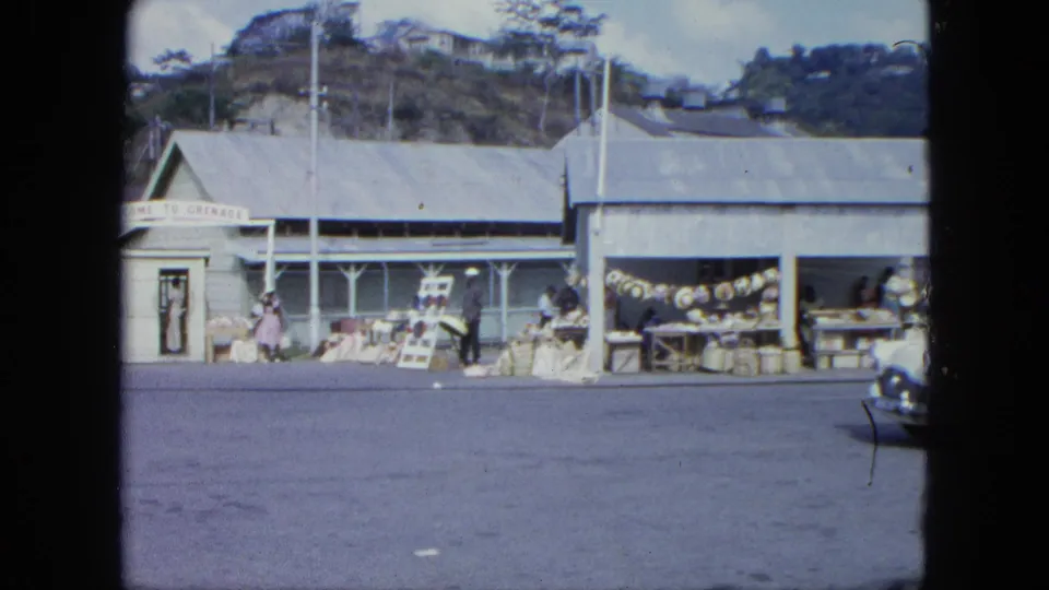 1960s Grenada market frontage with people browsing or selling goods beneath corrugated roofs and hills behind the street.