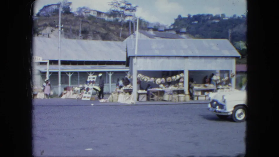 A white vintage car begins to pass a Grenada roadside market where people stand near open stalls and displayed goods.