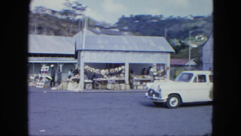 A white vintage car passes a 1960s Grenada roadside market with open stalls, displayed goods, and people under metal roofs.