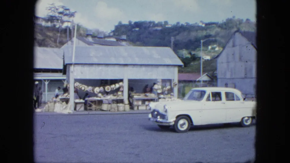 1964 8mm view of a white vintage car driving past a small roadside market with people and goods under a metal roof.