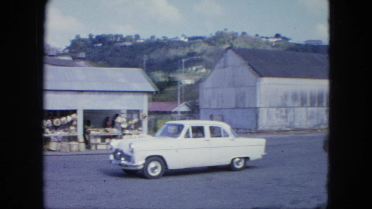 White sedan moving past market stalls with hillside and warehouse buildings behind.