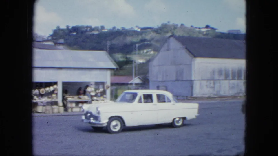 White mid-century sedan continues along a Grenada street past market goods and corrugated buildings, with green hills in the background.