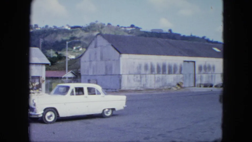 The white car exits frame beside a large weathered corrugated warehouse, with a hillside settlement visible above the road.