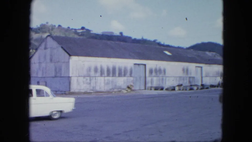 A white vintage car is nearly out of frame while the shot holds on a long corrugated warehouse, open street, and hillside beyond.