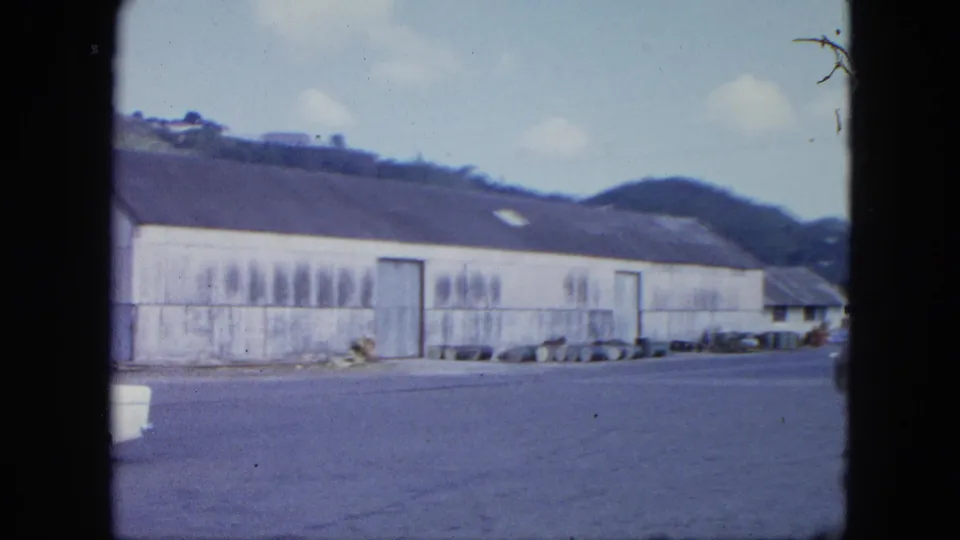 1964 home-movie frame showing a long weathered warehouse, open paved yard, parked items, and green hills beyond.