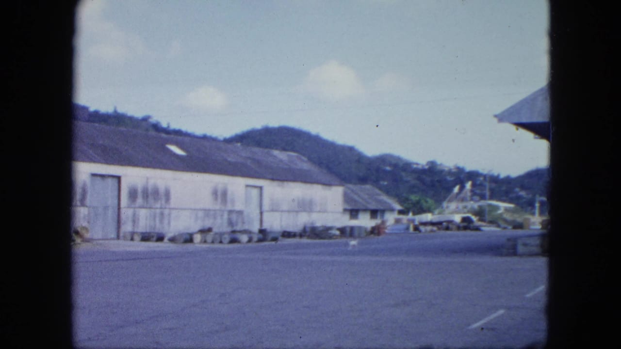 Industrial yard with a long warehouse, distant vehicles, hills, and a road area at right.