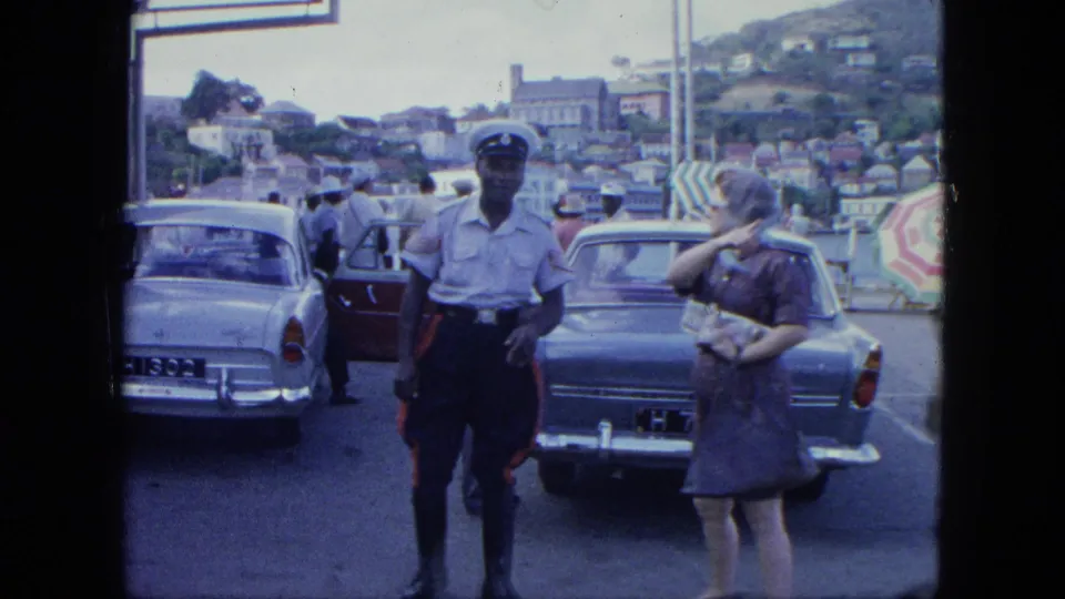 A uniformed guard stands near a woman in sunglasses beside classic cars, with hillside town buildings behind them.