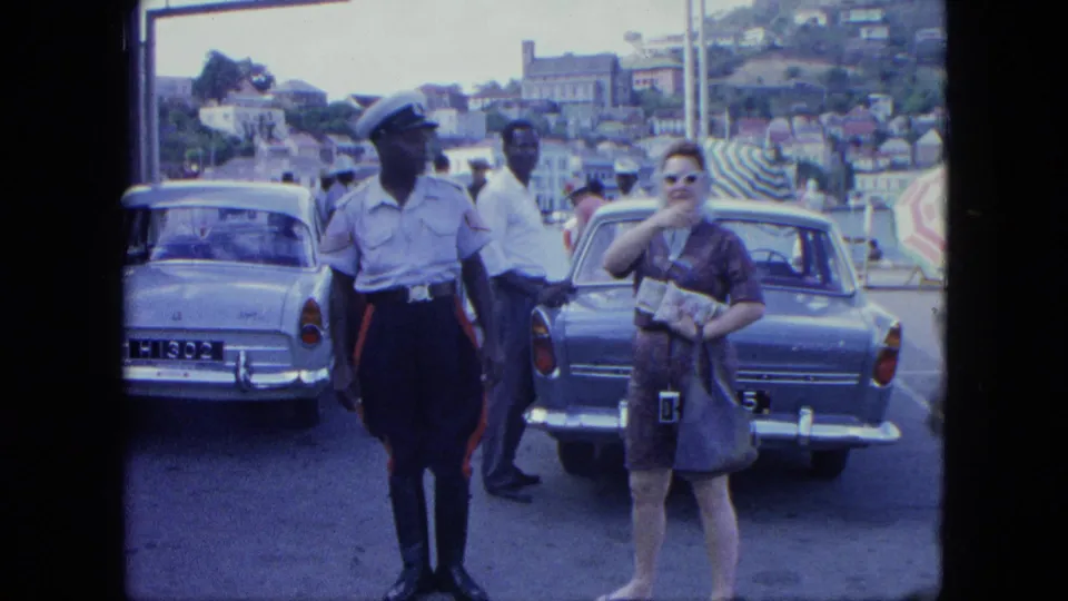 A guard, a woman with a handbag, and another man stand near classic cars, striped barriers, and hillside buildings.