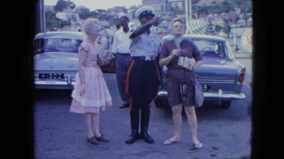 A uniformed officer points outward as two women stand nearby in a parking area, with vintage cars, pedestrians, and a hillside town in the background.
