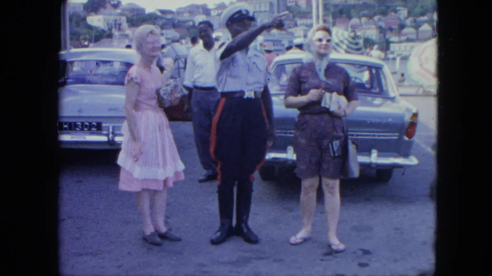 A police officer in a peaked cap gestures while two women stand beside him in a public lot with classic cars and hillside buildings behind them.