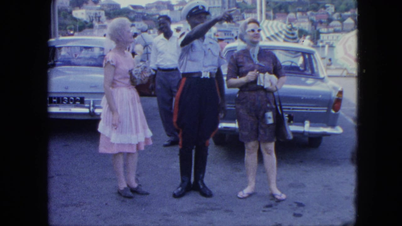 Tourists stand beside parked cars while a uniformed officer points in a street scene.