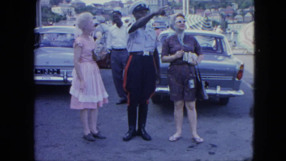1964 Grenada home-movie frame of tourists near classic cars speaking with a uniformed officer who points across the street.