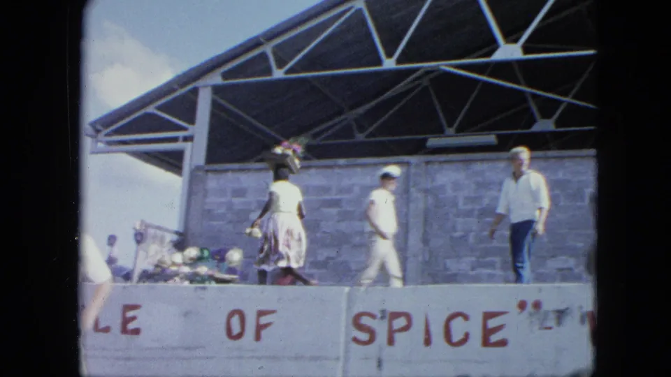A public stage or pavilion with people walking past a painted sign, filmed in the supplied 1964 Grenada travel footage.