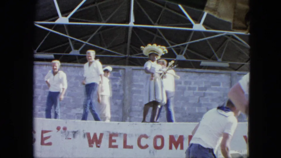 A performer in white dress and decorative hat stands on a public stage while several people watch or walk past.