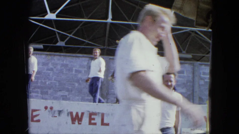 A foreground passerby partly obscures a pavilion stage where people stand behind a partially visible welcome sign.