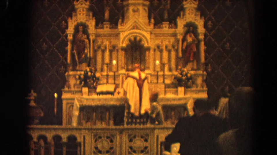 1964 Harvard, Illinois home-movie frame showing a priest at a carved Catholic altar with candles, statues, and congregants in shadow.