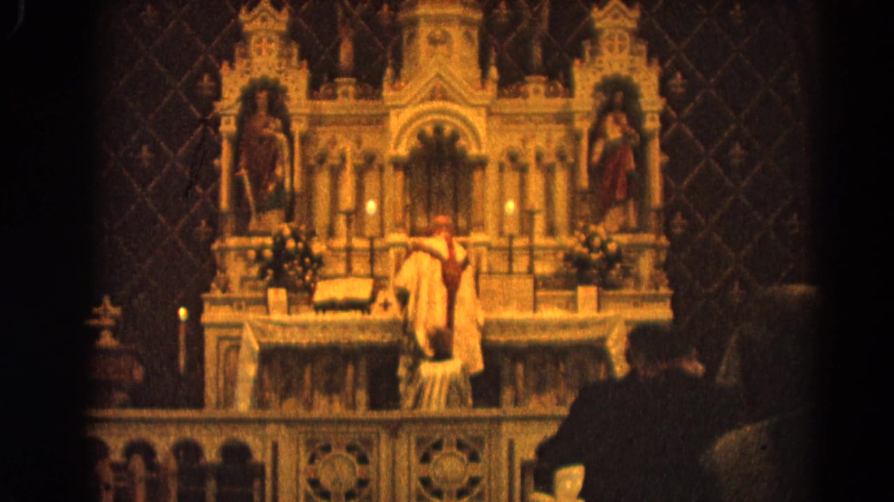 Priest at a Catholic altar in Harvard, Illinois, with warm candlelight, carved reredos, and seated congregants partly visible.