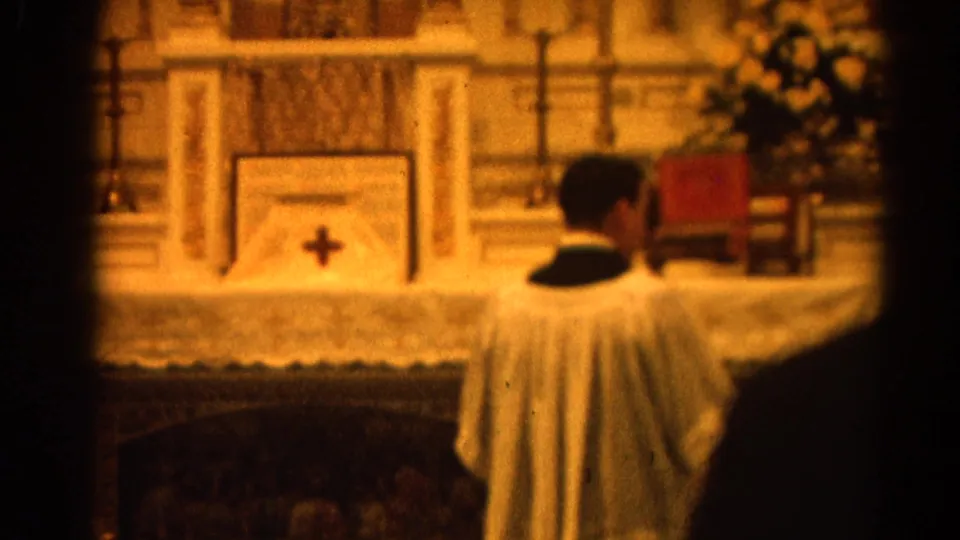 1964 Harvard, Illinois 8mm frame showing a clergy member facing a Catholic altar with altar cloth, crucifix, and candles.