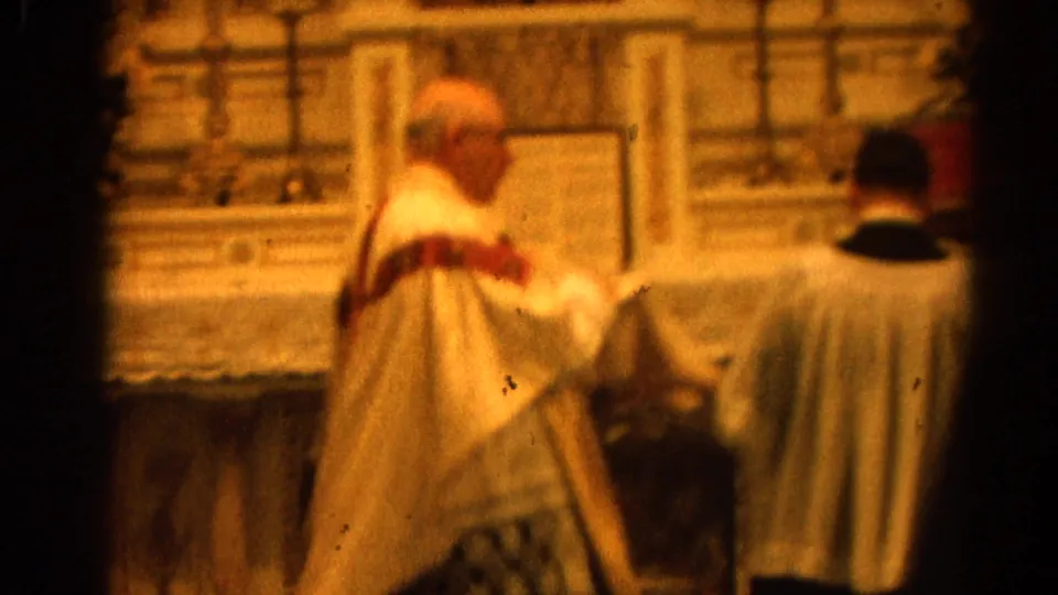 1964 8mm frame of a Catholic priest in white and red vestments beside an assisting clergy member at the altar.